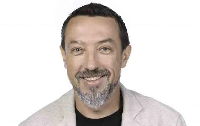 A smiling man with short dark hair and a gray beard, wearing a light-colored blazer over a black shirt, against a white background.