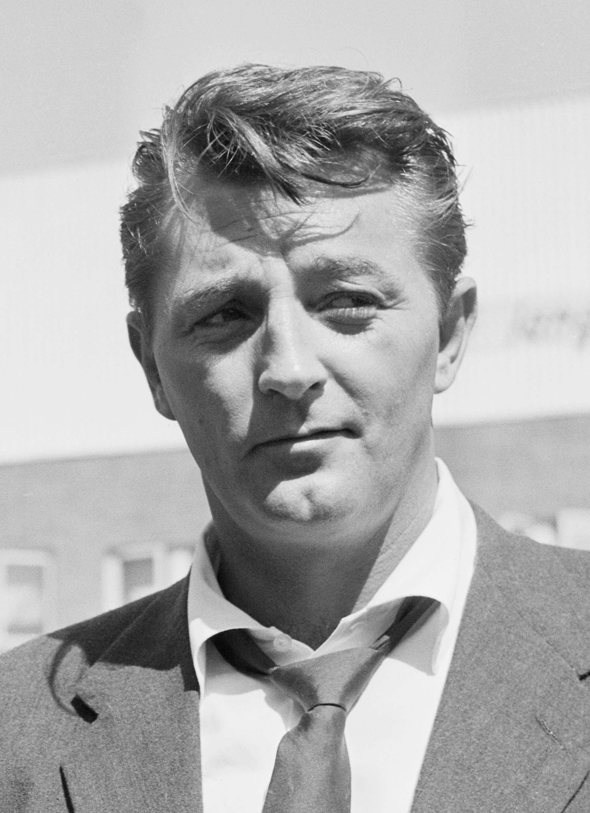 Black and white photo of a young man in a suit and tie, with wavy hair, outdoors, with sky and some distant structures in background.