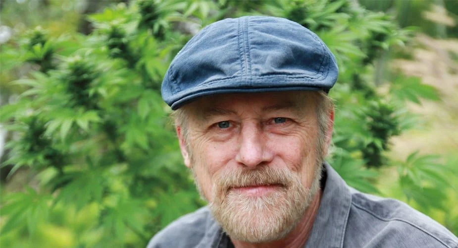 A middle-aged man with a beard and mustache wearing a blue cap and gray shirt, standing outdoors in front of green foliage.