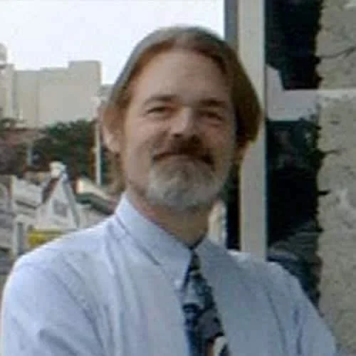A man with shoulder-length hair and a beard, wearing a light blue dress shirt and a patterned tie, standing outdoors with buildings and trees in the background.