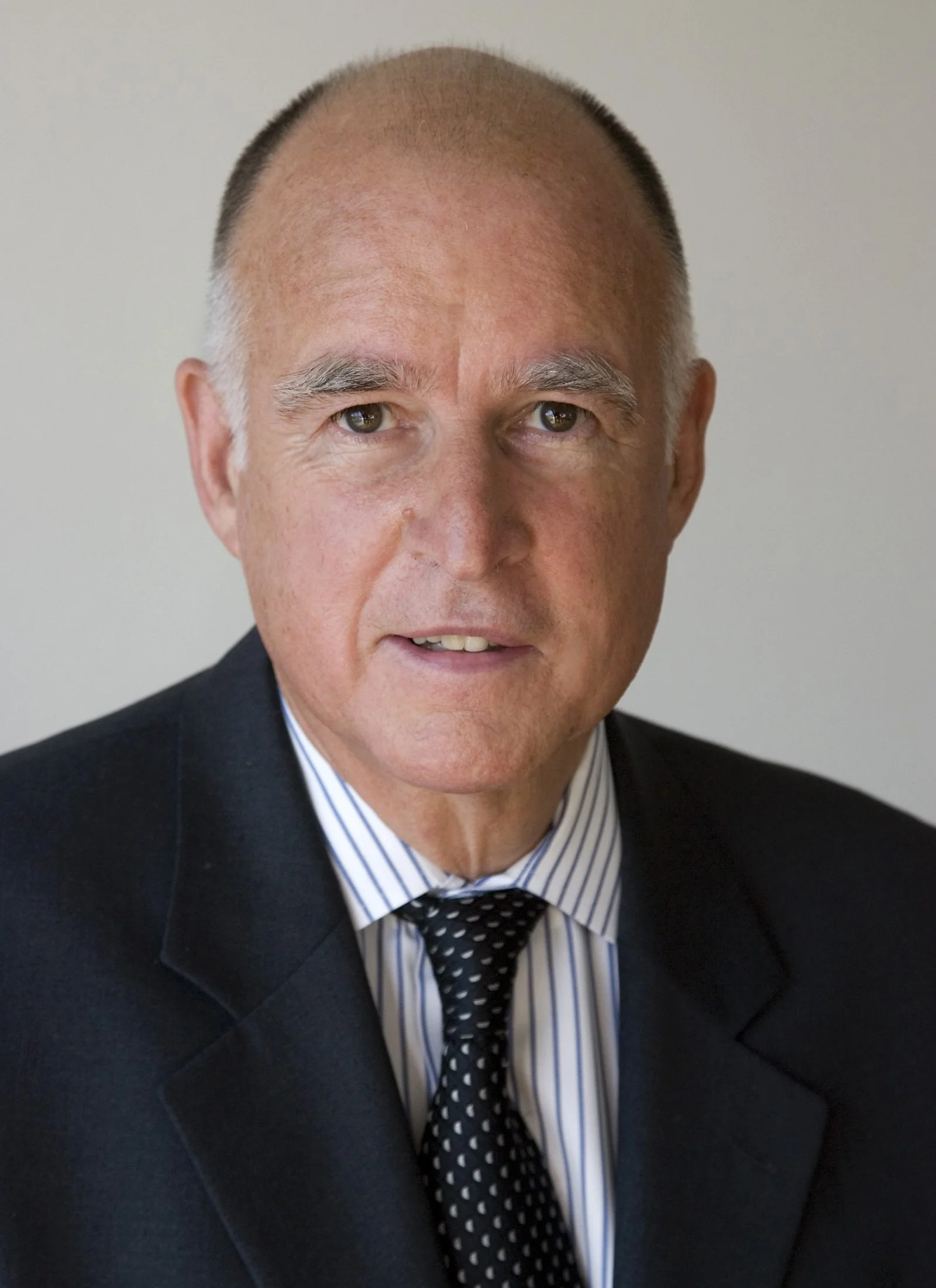 Close-up of a middle-aged man with a receding hairline, wearing a dark suit, striped shirt, and polka dot tie, against a plain background.