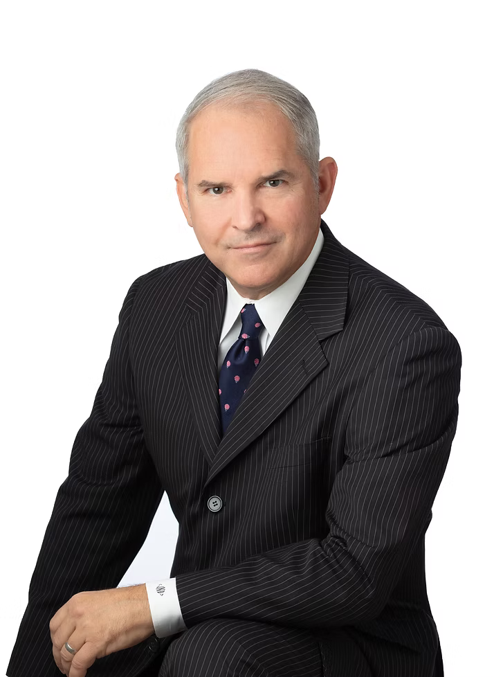 A middle-aged man in a black pinstripe suit, white shirt, and dark tie with small pink and white pattern, posing against a white background.