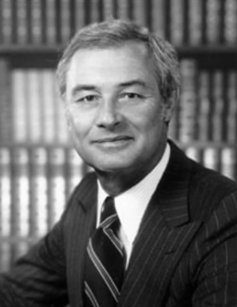 Black and white photo of a middle-aged man in a suit with a tie, sitting in front of a bookshelf filled with books.