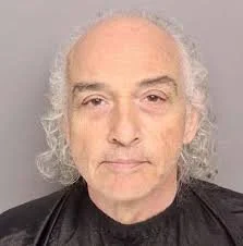 Close-up portrait of an older man with white, curly hair and a serious expression, against a plain gray background.