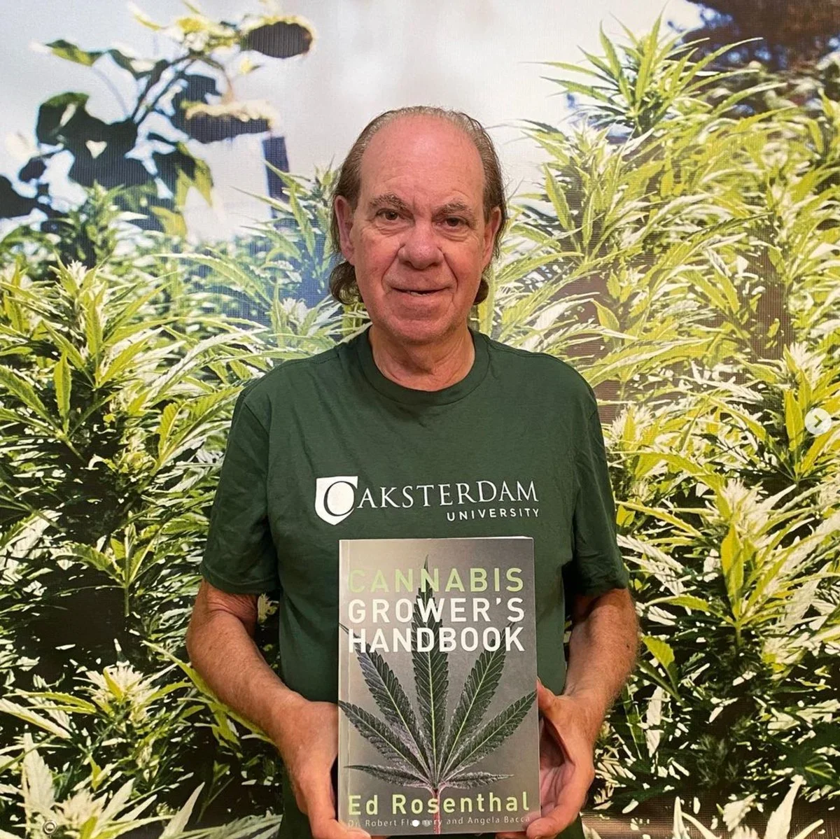 A man in a green 'Akademdam University' t-shirt standing in front of cannabis plants, holding a book titled 'Cannabis Grower's Handbook' by Ed Rosenthal.