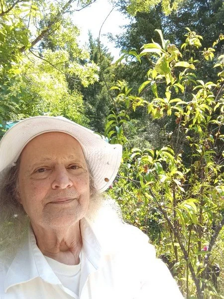 Older man with long gray hair wearing a white hat and white shirt outdoors among green trees and plants, taking a selfie on a sunny day.