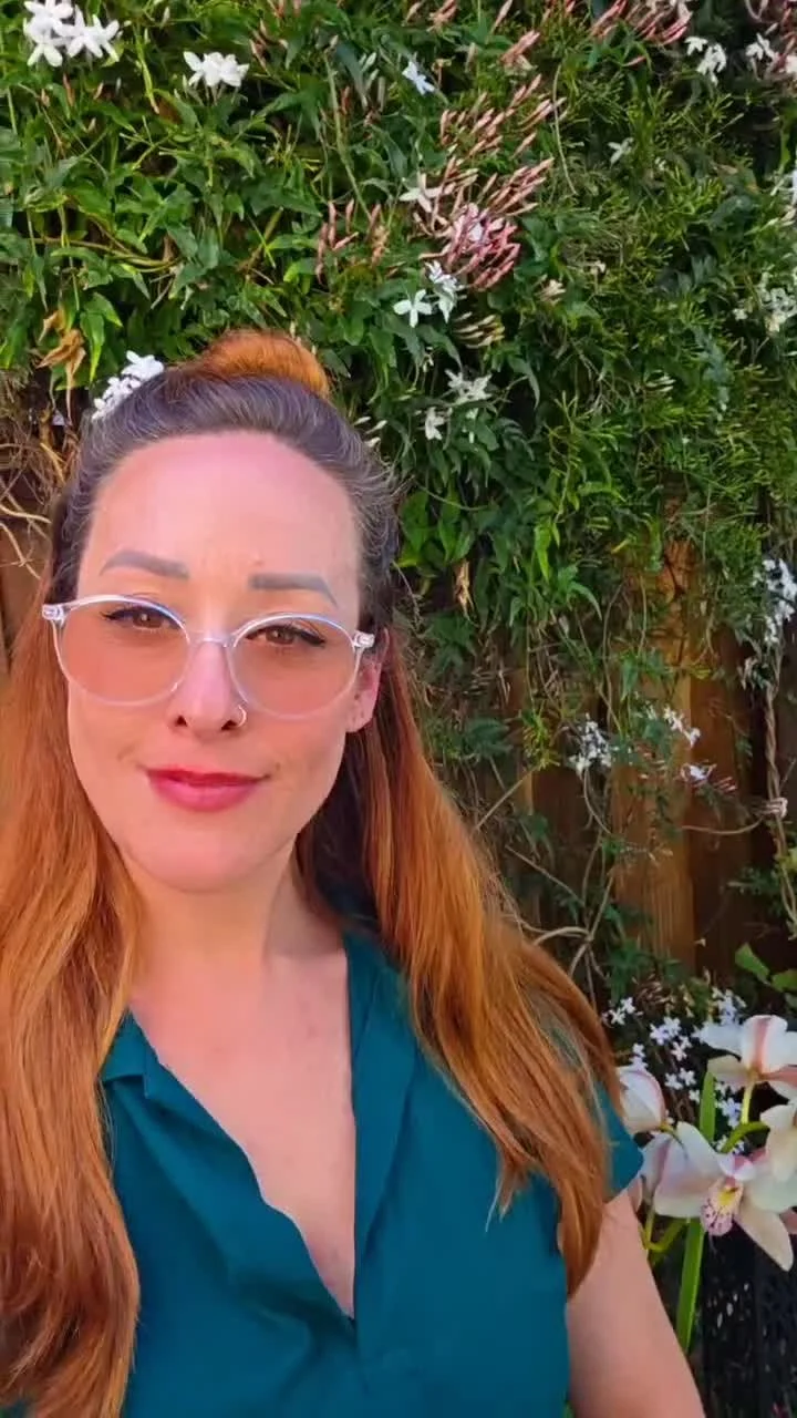 A woman with long red hair, wearing glasses and a teal top, taking a selfie outdoors in front of a lush green background with white and pink flowers.