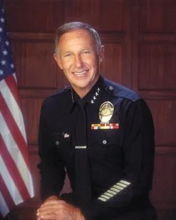 A smiling police officer in uniform standing in front of an American flag and wooden paneling.