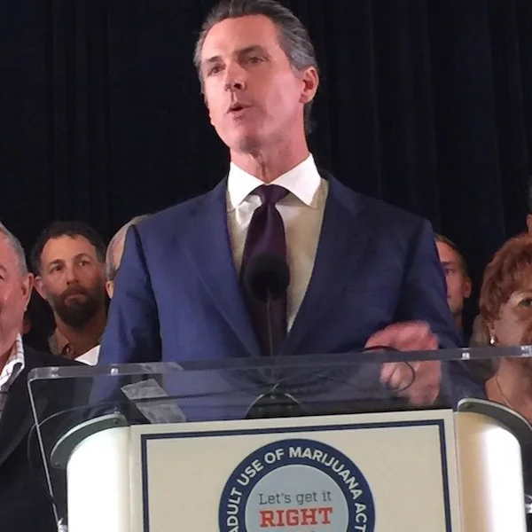 A man in a blue suit and white shirt speaking at a podium, with a group of people behind him, in front of a dark curtain.