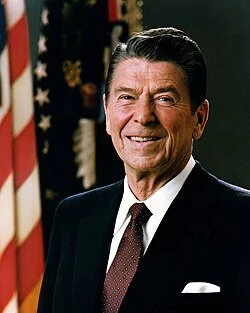 A smiling man in a suit standing in front of American flags.