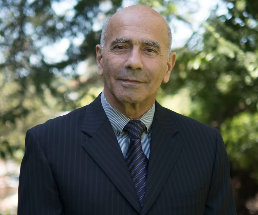 An older man in a pinstriped suit with a striped tie, standing outdoors with green trees in the background.