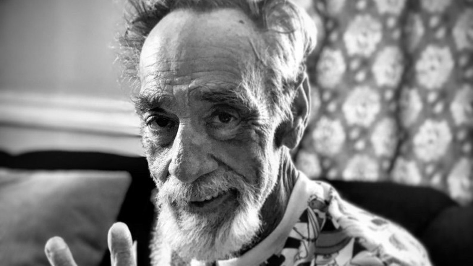 Close-up, black-and-white photo of an elderly man with white hair and beard, smiling slightly, in front of a patterned background.