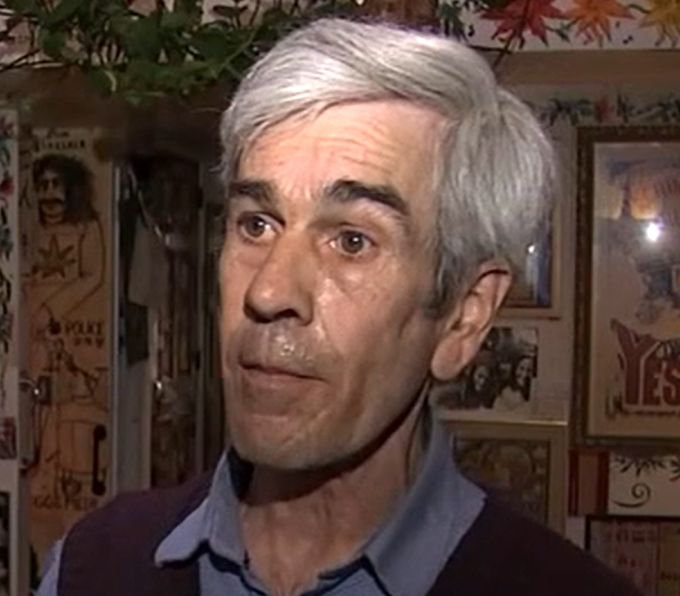 An elderly man with white hair, wearing a collared shirt and vest, talking in a room filled with vintage posters and framed photos.