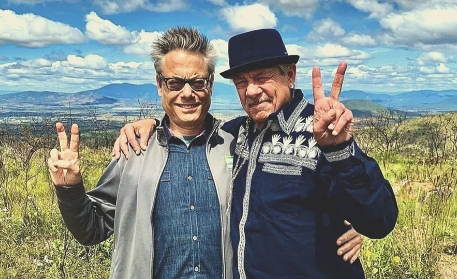 Two men standing outdoors in a field, smiling and making peace signs with their hands, with a scenic landscape and cloudy sky in the background.