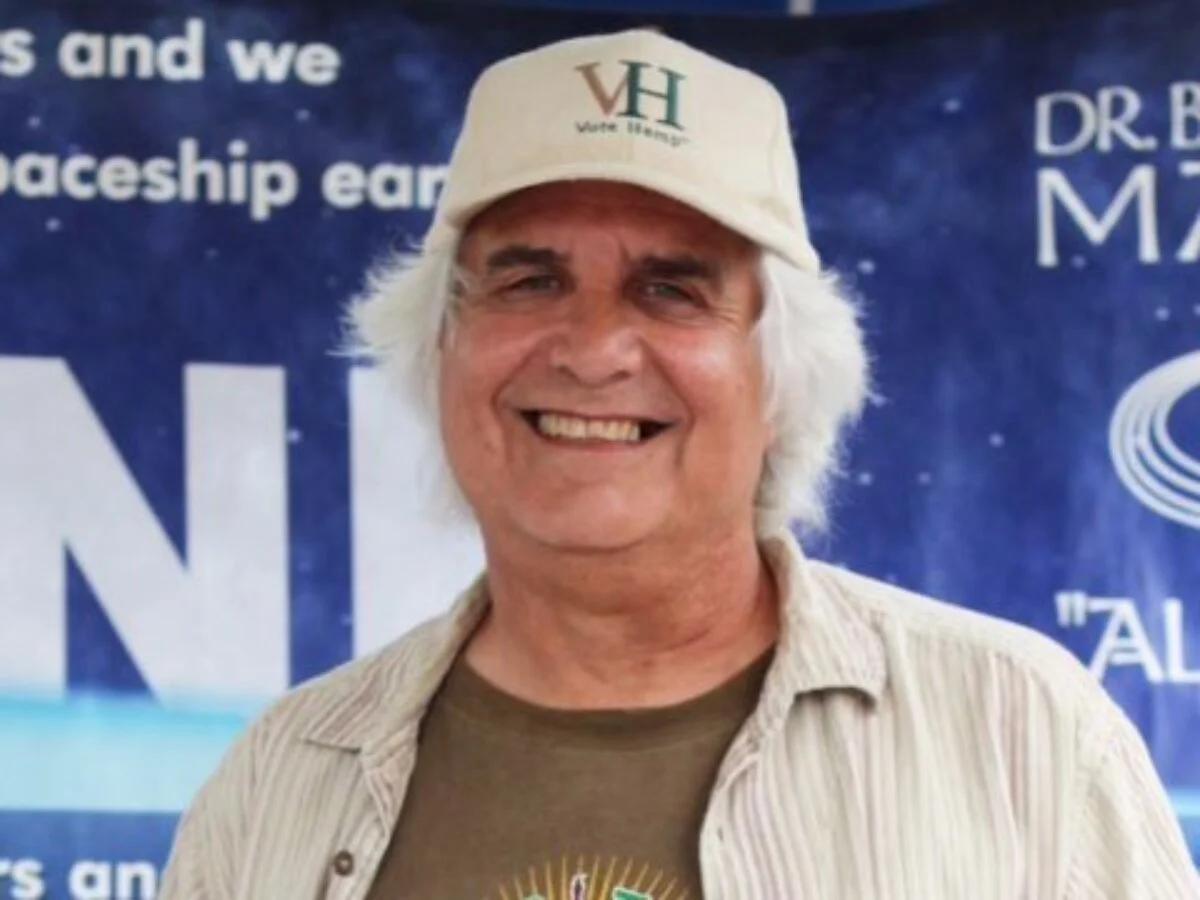 A smiling man with gray hair wearing a beige baseball cap and a beige button-down shirt, standing in front of a blue space-themed backdrop.