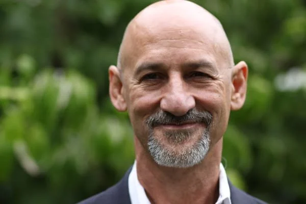 Close-up of a bald man with a gray goatee, smiling outdoors with green foliage in the background.