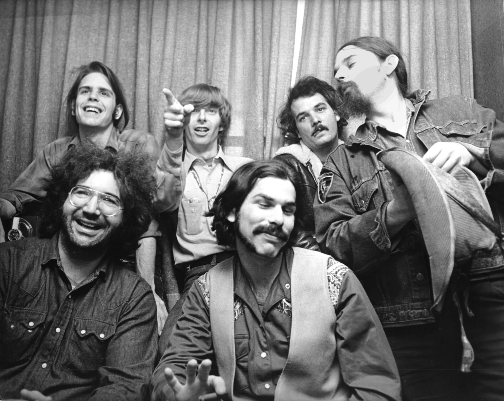 A black and white photo of a group of six men, some with long hair and beards, posing together in front of curtains. They appear relaxed and happy, with some smiling and making gestures.