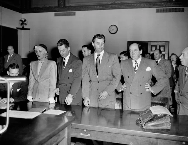Black and white photo of men and women in formal attire standing in a courtroom or meeting room with a large table, bookshelves, and a clock on the wall.