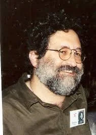 A man with curly dark hair and glasses, smiling with a beard and mustache, wearing a dark shirt and a badge, standing outdoors at night.