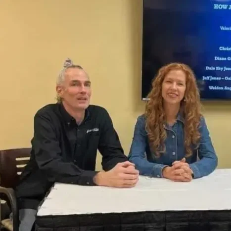 A man and a woman sitting at a table in a room with a yellow wall and a large screen behind them. The man has gray hair tied in a bun, wears a black shirt, and is looking to the left. The woman has long, curly red hair, wears a denim jacket, and is smiling. They are sitting in front of a white tablecloth.
