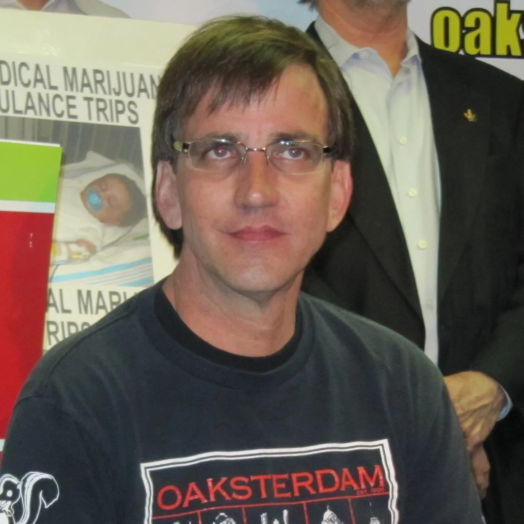 A man with glasses and dark hair wearing a black T-shirt with red and white text that says 'OAKSTEDAM'. He is in front of a poster about medical marijuana ambulance trips, with other people partially visible in the background.