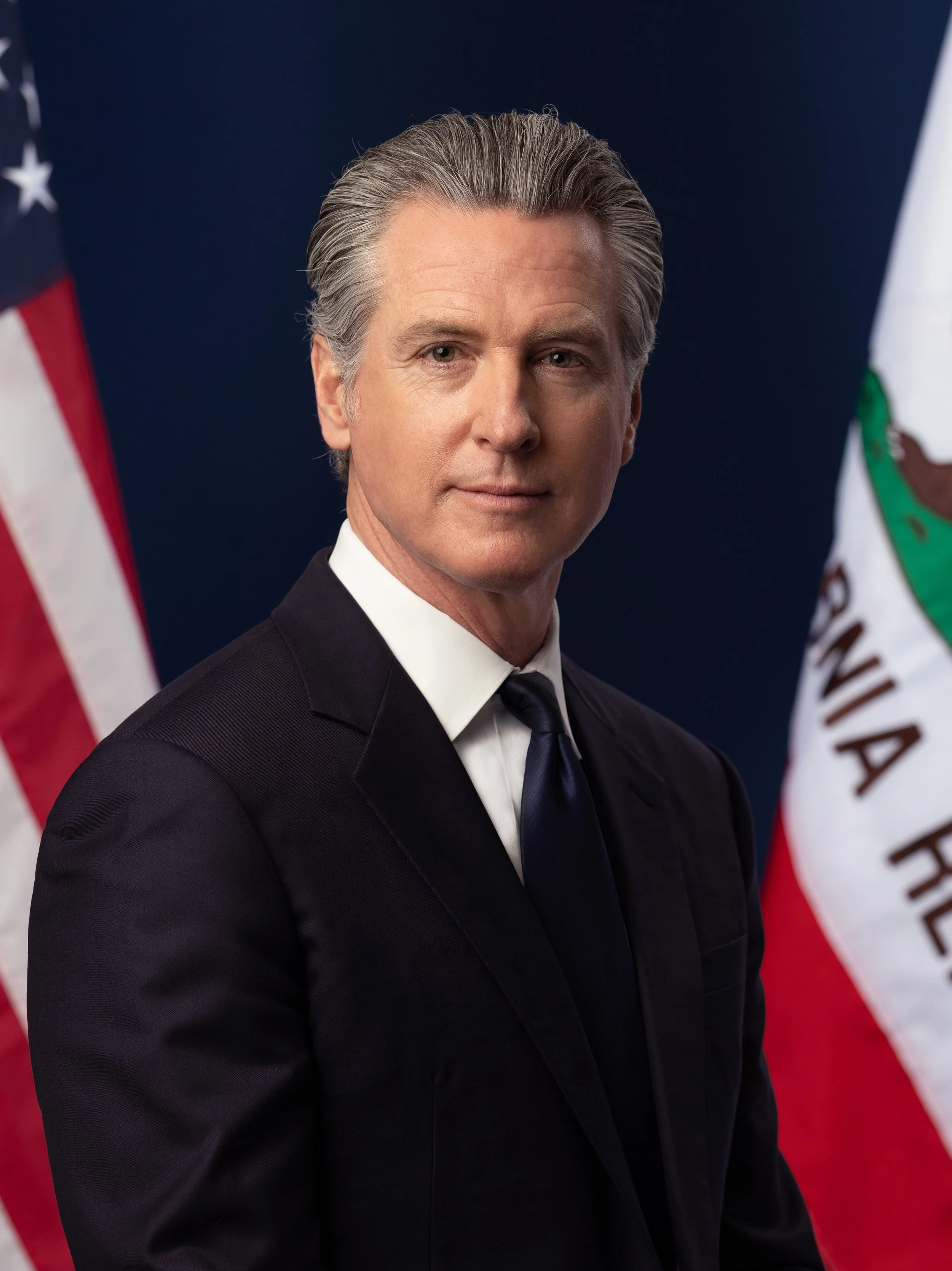 A man in a dark suit and tie in front of American and California flags.