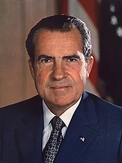 A formal portrait of a middle-aged man with dark hair, wearing a navy suit, white shirt, and patterned tie, standing in front of an American flag.
