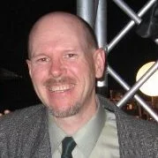 A smiling man with a beard and short hair, wearing a gray jacket and a tie, standing in front of a metallic background.