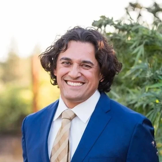 Portrait of a smiling man in a blue suit and beige tie outdoors with greenery in the background.