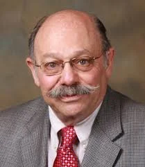 Portrait of an older man with glasses, a mustache, wearing a gray blazer, white shirt, and red tie, against a plain background.