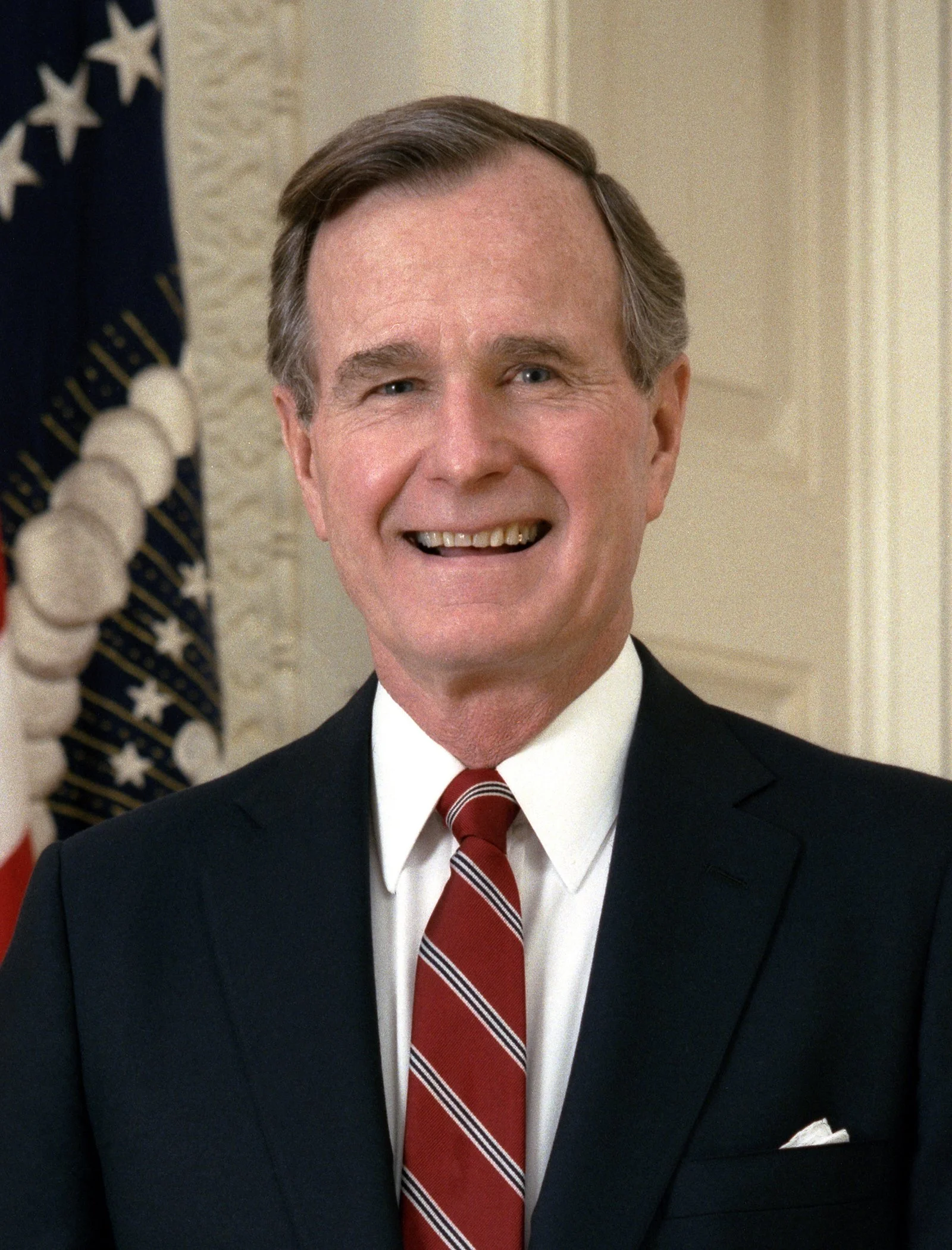 Close-up of a smiling man in a suit and striped tie, with part of an American flag in the background.