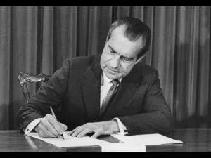 A man in a suit sitting at a desk, signing a document with a pen.