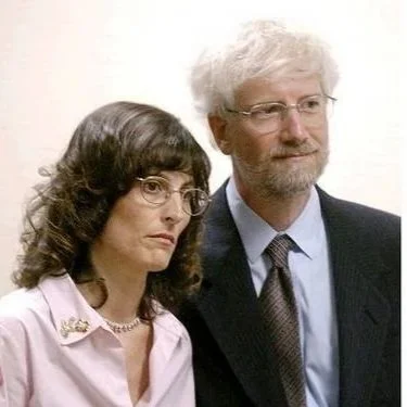 A woman with dark, curly hair and glasses, and a man with white hair and glasses, both dressed in professional attire, standing together.
