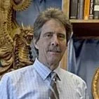 A middle-aged man with curly hair and a beard, wearing a dress shirt and tie, standing in an indoor setting with shelves and decorative items in the background.