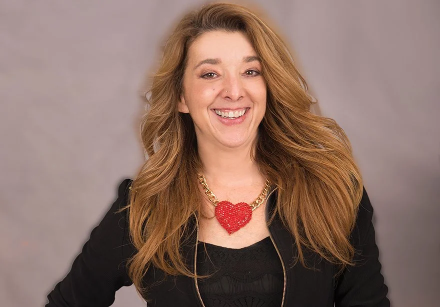 A smiling woman with long, wavy red hair wearing a black top and a chunky gold chain necklace with a large red heart pendant.