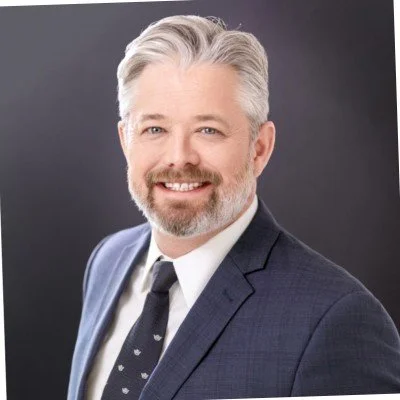 Professional middle-aged man with gray hair and beard smiling in a dark suit and tie against a dark background.