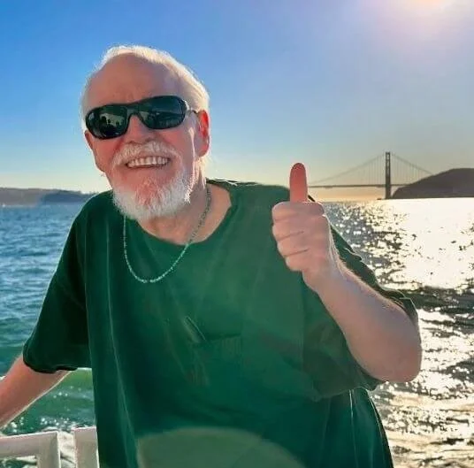 Older man smiling with sunglasses, giving a thumbs up, on a boat with water and a bridge in the background during sunset.