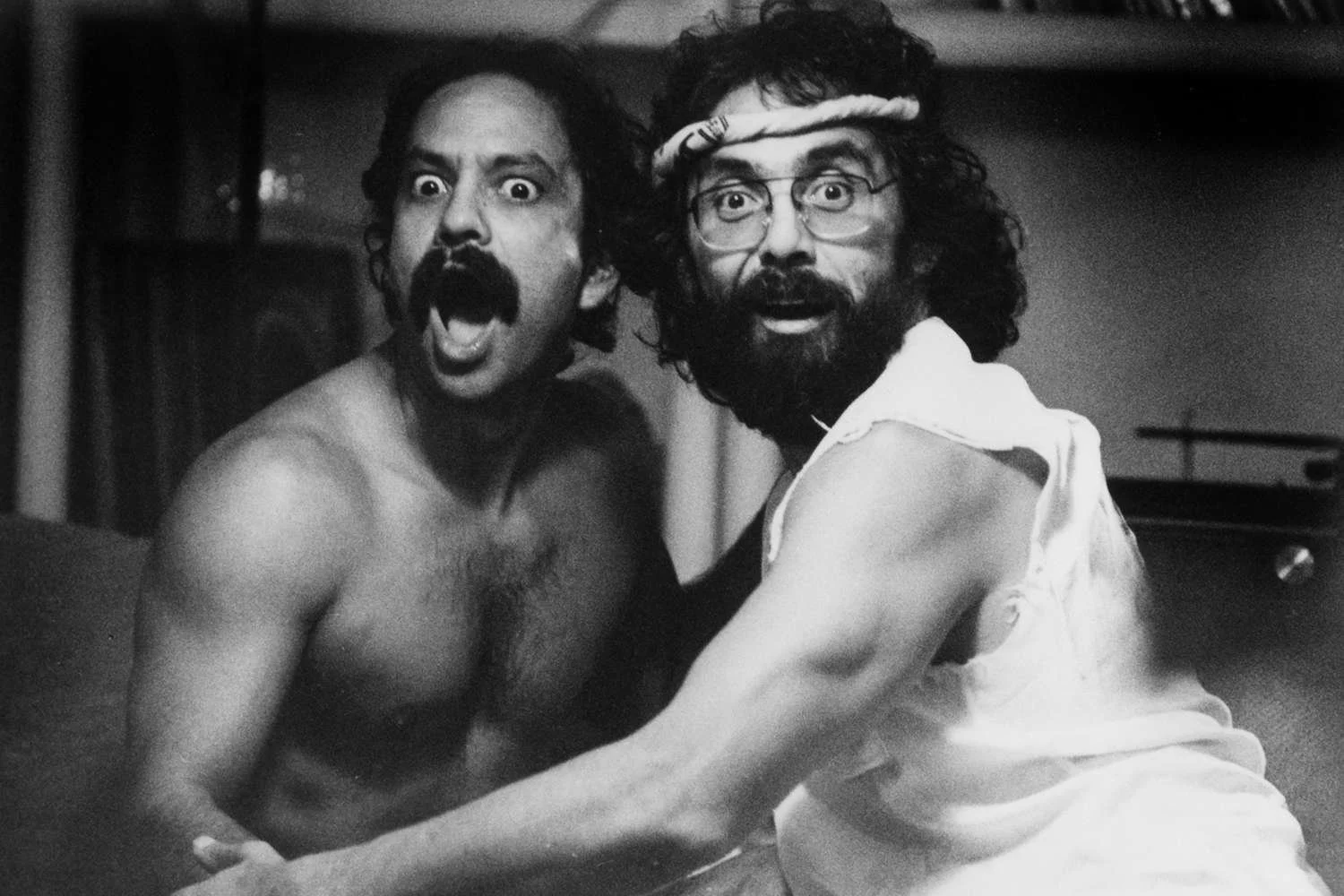 Black and white image of two men with surprised or excited expressions, one shirtless with a mustache and the other wearing glasses, a headband, and a sleeveless shirt.