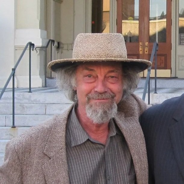 A man with gray hair and beard wearing a fedora hat, a gray pinstripe shirt, and a brown tweed blazer, standing outdoors in front of a building with stairs and a railing.