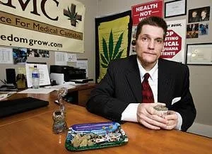 Man in a suit sitting at a desk with a marijuana leaf poster in the background, holding a glass of water.