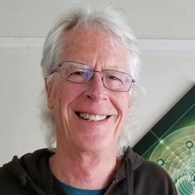 A smiling elderly man with white hair and glasses, wearing a dark hoodie, standing in front of a green and black abstract background.