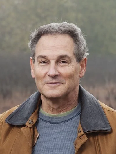 A middle-aged man with gray hair, wearing a brown jacket over a blue shirt, standing outdoors with a blurred natural background.