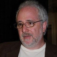 Close-up of a middle-aged man with gray hair, beard, glasses, wearing a dark jacket and white shirt, indoors.