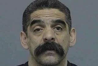 Close-up of a man with dark hair, thick mustache, and eyebrows, looking directly at the camera against a plain background.