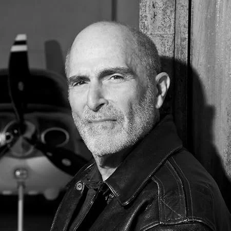 Black and white photo of a middle-aged man with a beard, wearing a leather jacket, leaning against a wooden wall.
