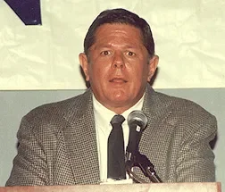 A man in a gray blazer and white shirt speaking at a microphone during a formal event or press conference.