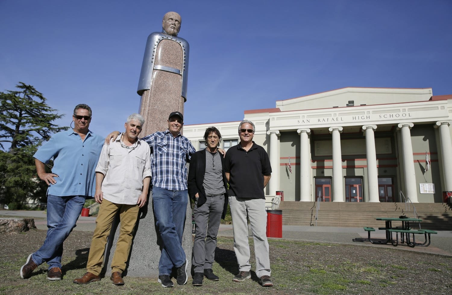 Five men standing in front of San Rafael High School with a large statue of a man behind them and trees nearby.