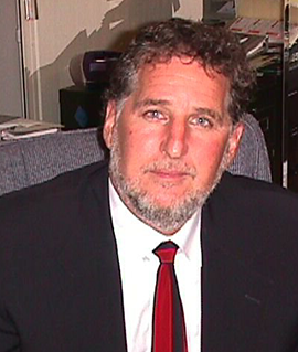 A man with curly hair, a beard, and blue eyes in a black suit, white shirt, and red tie sitting in an office with books and papers.