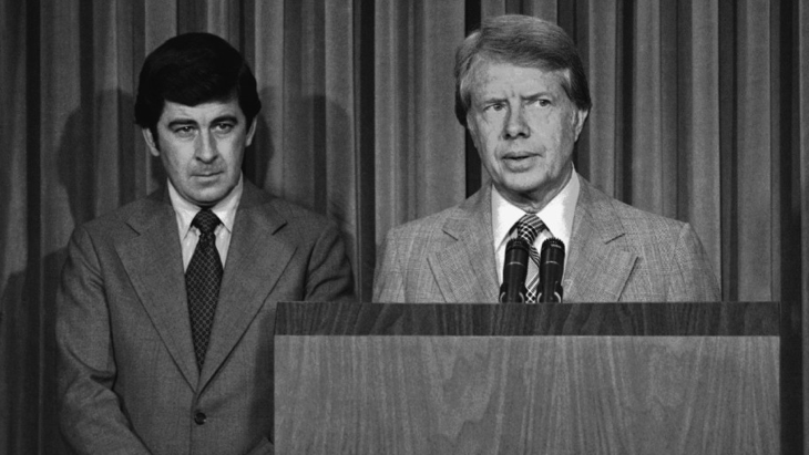 Two men in suits standing at a podium with microphones, with a curtain backdrop, during a formal event or press conference.