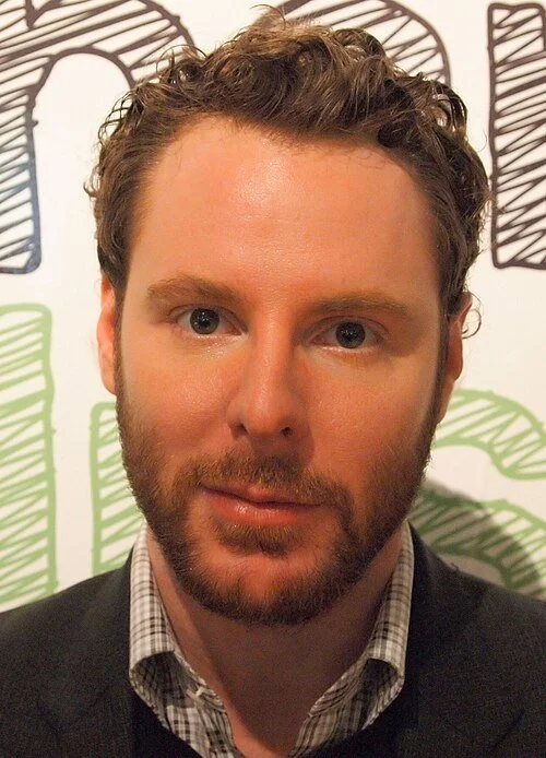 A man with short, curly brown hair and a beard, wearing a collared shirt and dark sweater, standing in front of a background with doodle-style drawings of trees.